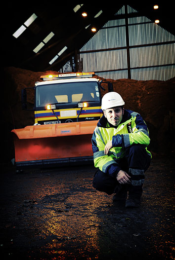 Workman in hard hat and high visibility jacket poses in front of a snow plough